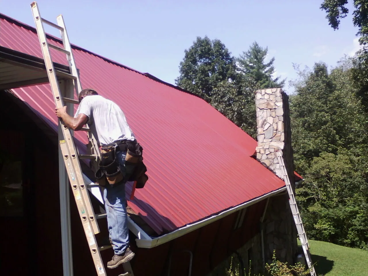 Roofer on a ladder beside a red metal roof and stone chimney — Soffit & Fascia Repair by Roofing Brothers in Dallas City